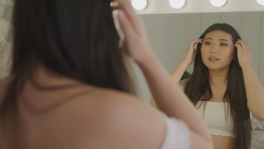 Asian woman with long hair and a white dress looks in the mirror and tidies her hair smilingly in the dressing room