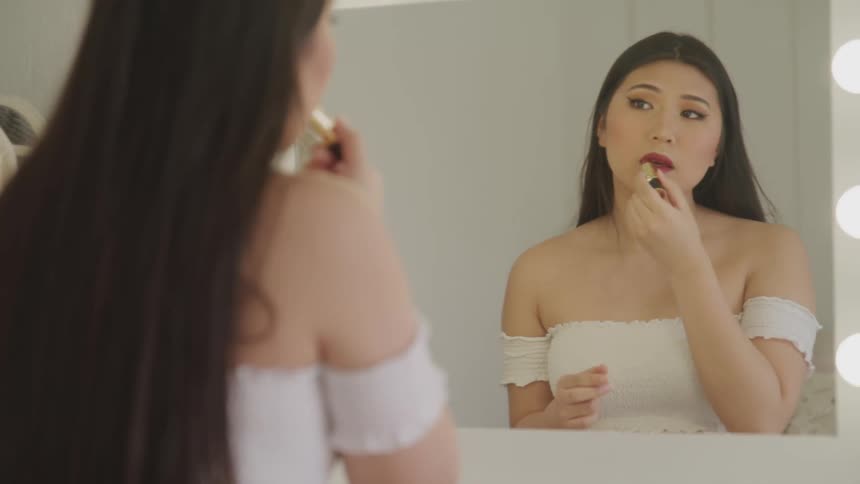 An asian woman applies red lipstick to her lips while sitting at a vanity