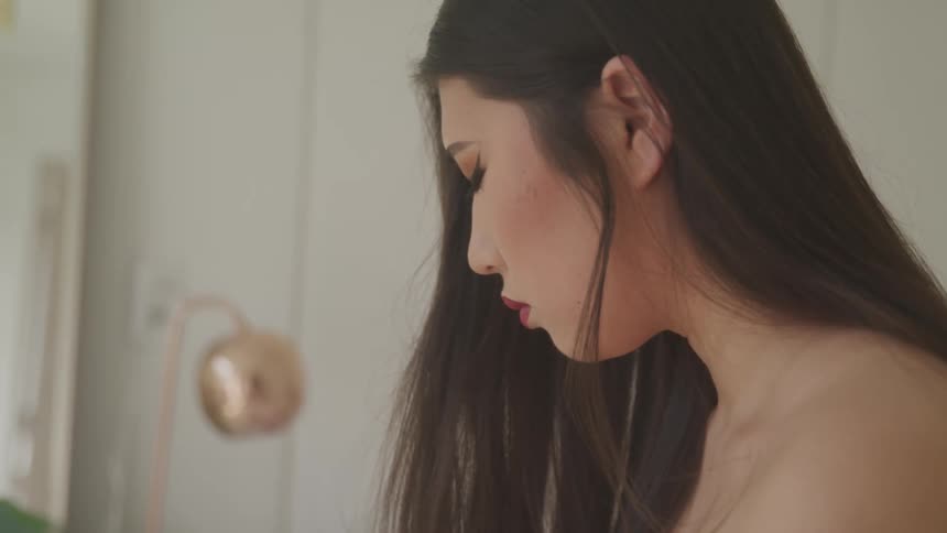 A young woman stands shirtless in her bedroom with her head bowed down and eyes closed