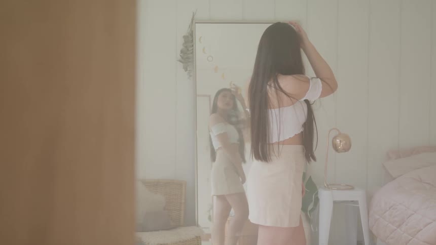 Woman primps and finger adjust her waist length hair while looking in mirror next to her bed