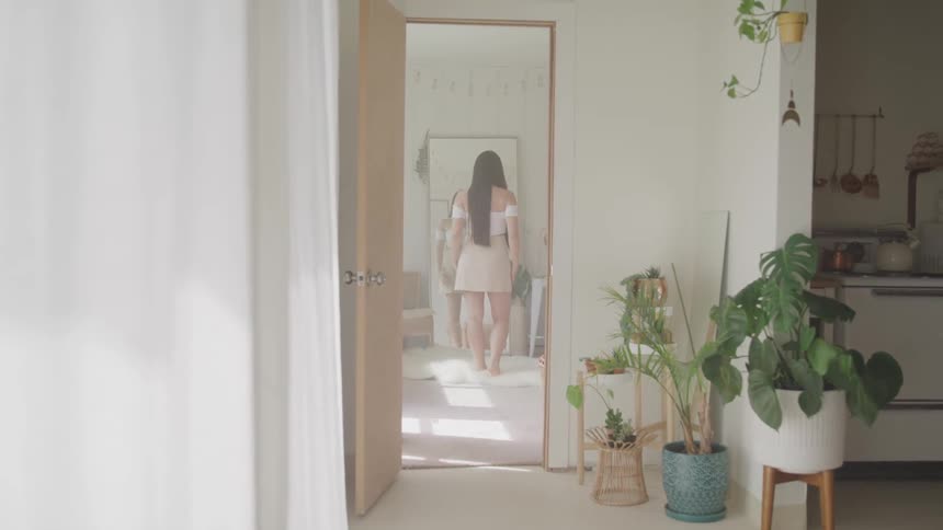 A woman in a white blouse and khaki skirt checks her outfit and hair in a floor length mirror in her apartment