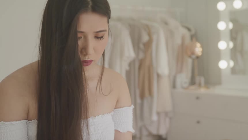 A woman in a white off the shoulder blouse sits in her all white bedroom with her head bowed and eyes closed