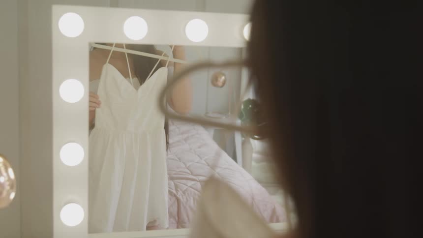 A young woman in her bedroom holds a dress against herself and looks in a mirror