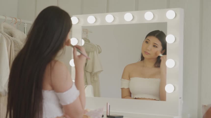 A young woman uses a make up brush to apply make up to her face in front of a large mirror