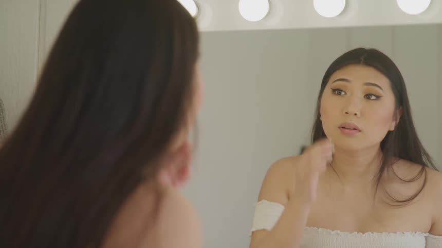 A woman standing in front of a mirror applying makeup on her face with a brush