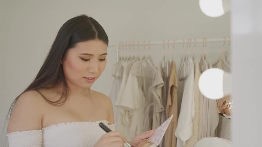 A young asian woman applies makeup to her face while looking into the mirror of a vanity