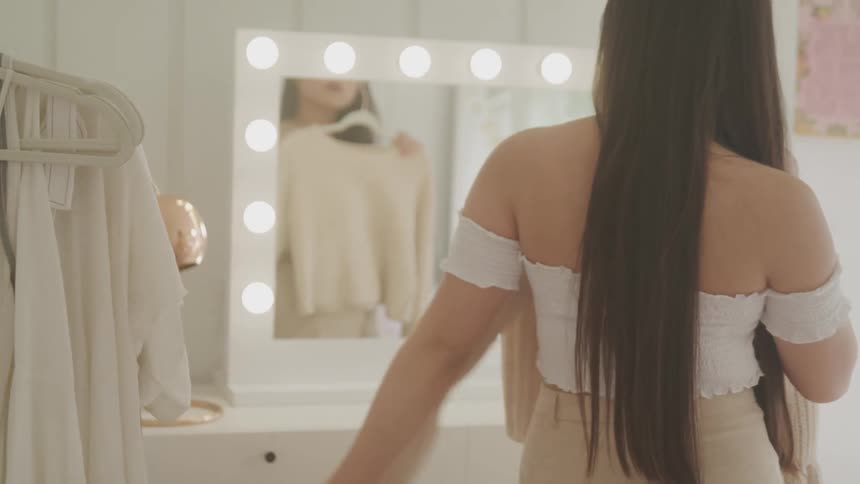 An asian woman holds up an article of clothing to herself and looks in the mirror