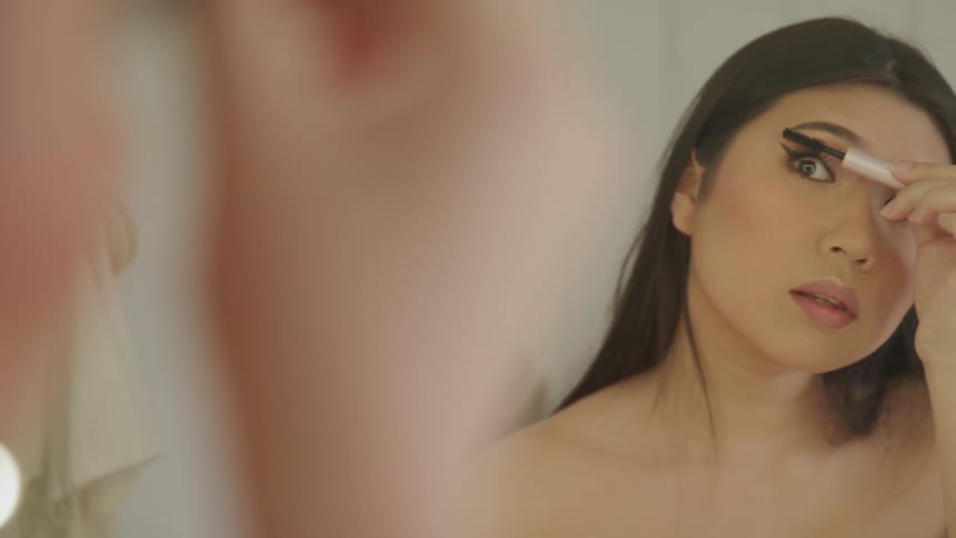 An asian female looks into a mirror while applying makeup to her eyelashes