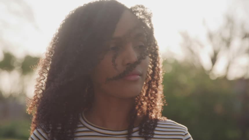 Black woman with curly hair and a sweatshirt faces the camera without eye contact as the sun shines in the background