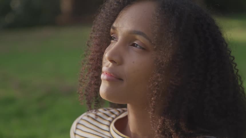 Side view of a black woman with curly hair standing on green grass and looking up at the sky smilingly