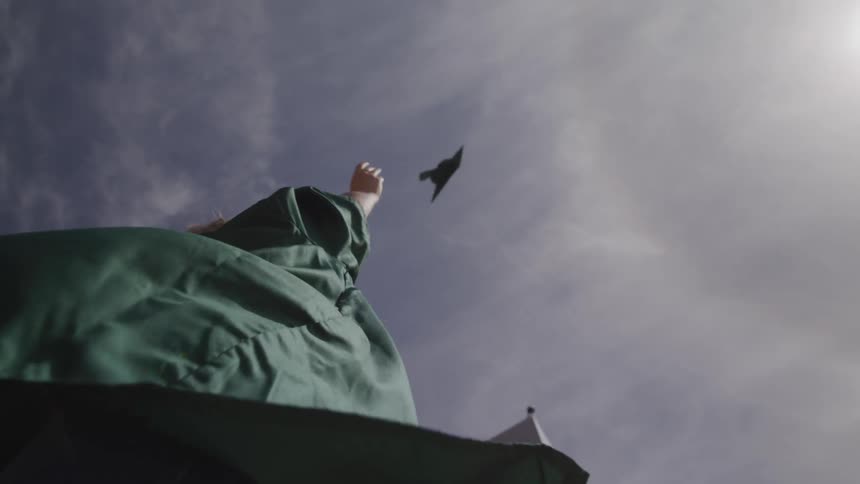 A woman wearing a green graduation gown jumps and tosses her cap in the air