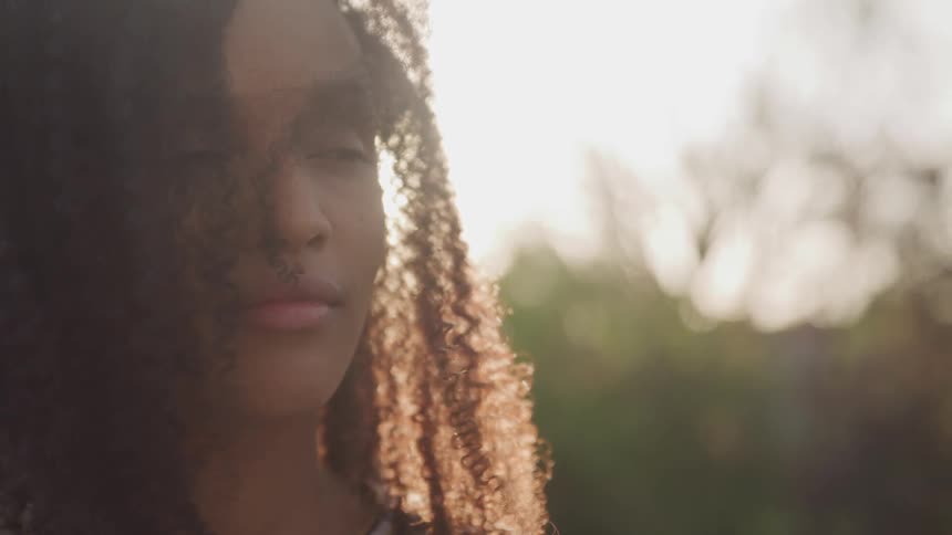 Black woman with curly hair smiles slightly as the sun shines slightly in the background
