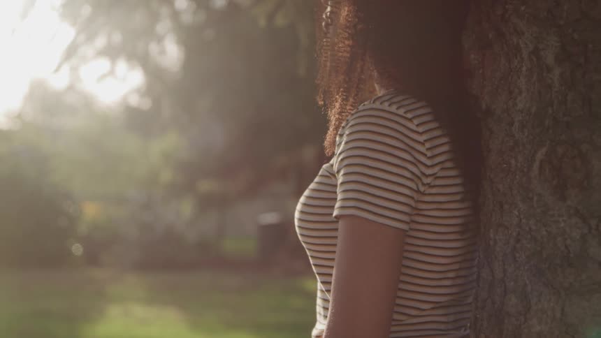 A woman looks up at the daytime sky while leaning against the trunk of a tree