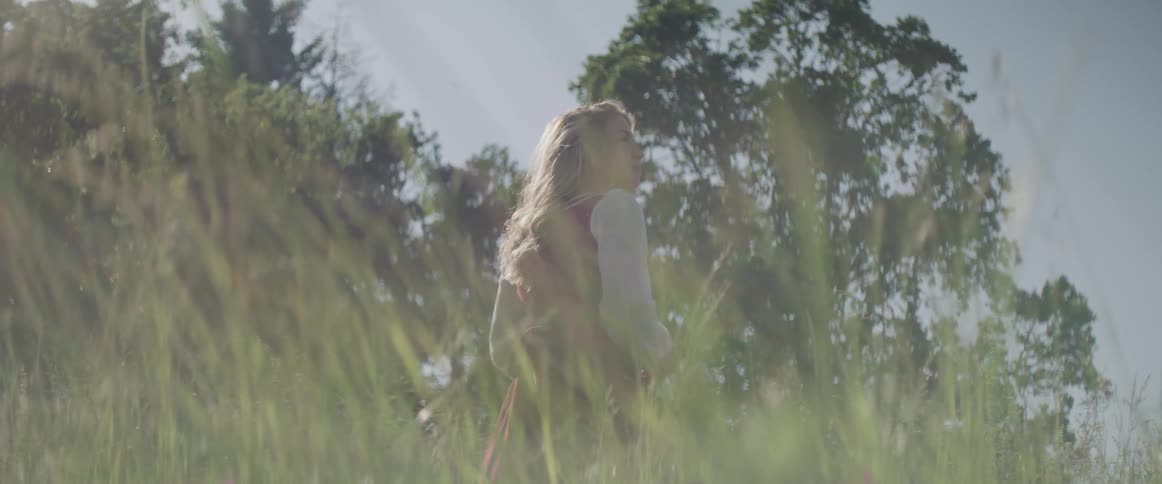 Woman with red dress stands in tall green grass and fiddles with her blonde hair