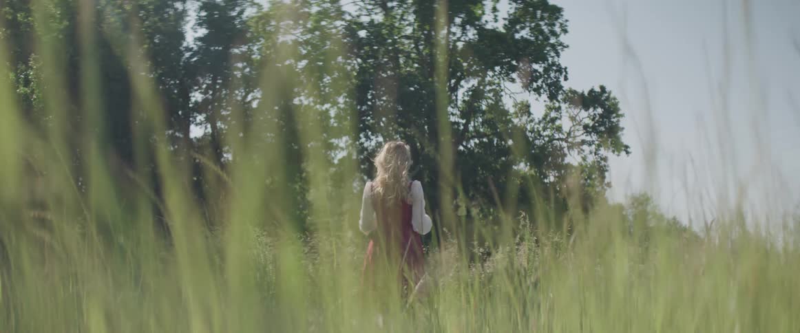 Woman with red dress stands in tall green grass and plays with her blonde hair
