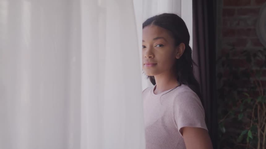 Girl with long hair and a pink t shirt stands next the windows and white curtains