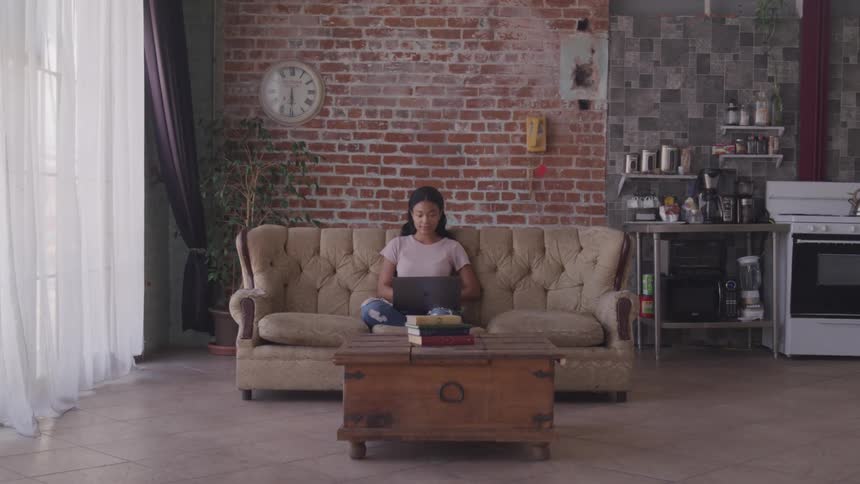 A woman sitting on a couch typing on a laptop with a wall in the background