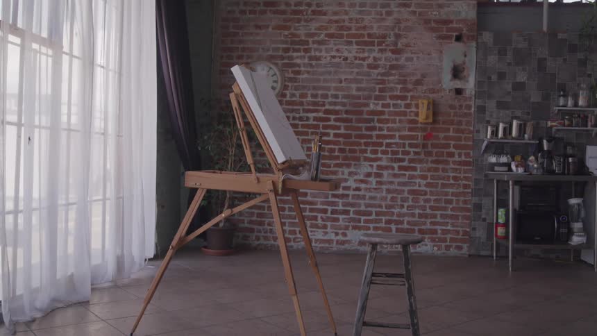 Artist easel and stool sit near curtained window in open area of room near kitchen