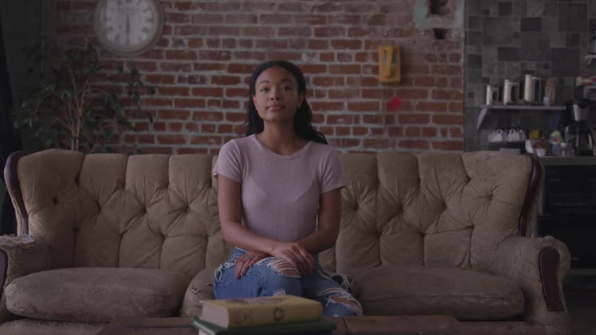 A young black woman sitting on a sofa turns her head to smile into the camera