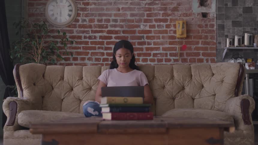 A woman sits on a couch with her legs folded under her working on a laptop