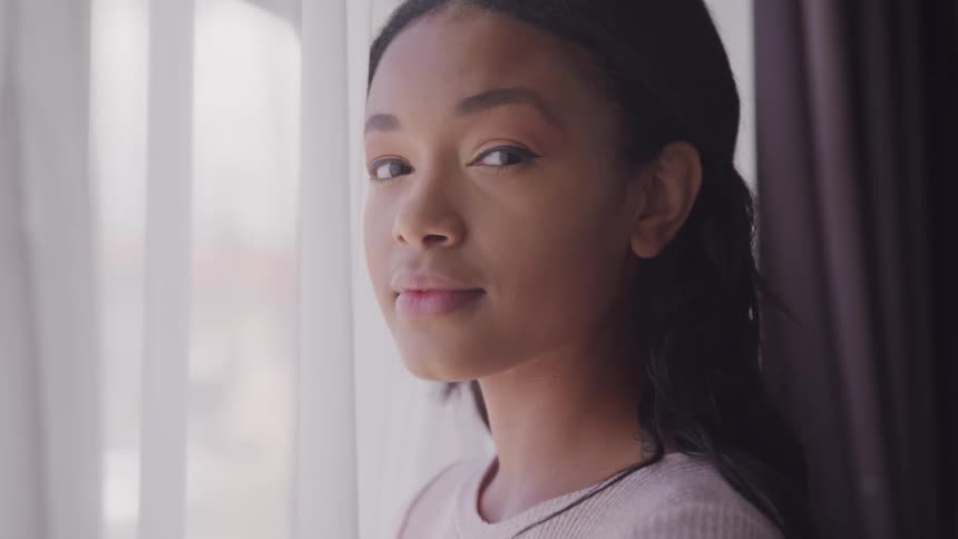 A teenager poses next to an open window as wind blows a sheer white curtain against her face
