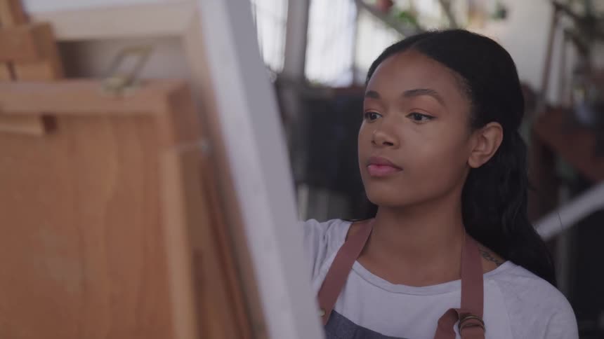 A young woman in an apron paints on a canvas being held by an easel
