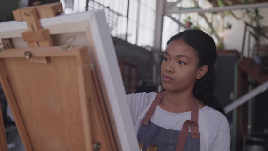 A woman with a apron sitting in front of a canvas and brushes