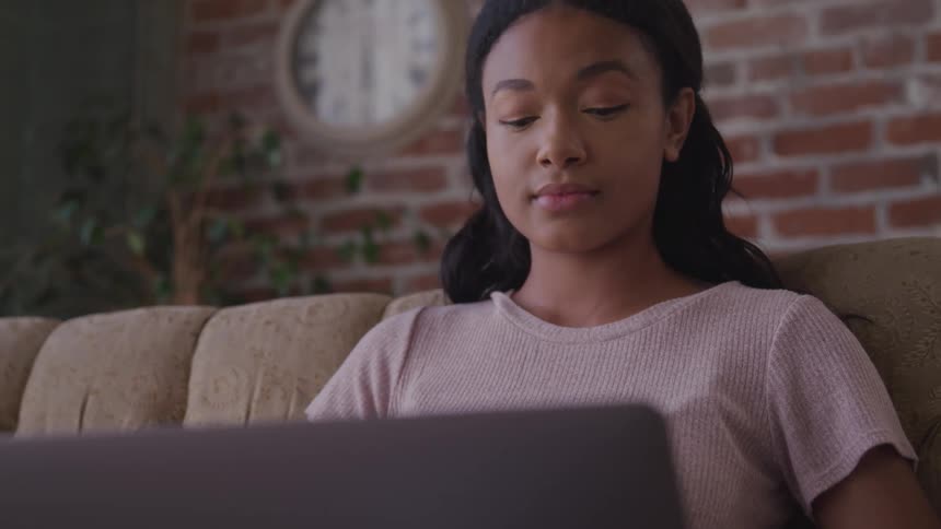 Woman with long hair and a pink t shirt sits on a sofa and uses a grey laptop in front of a brick wall