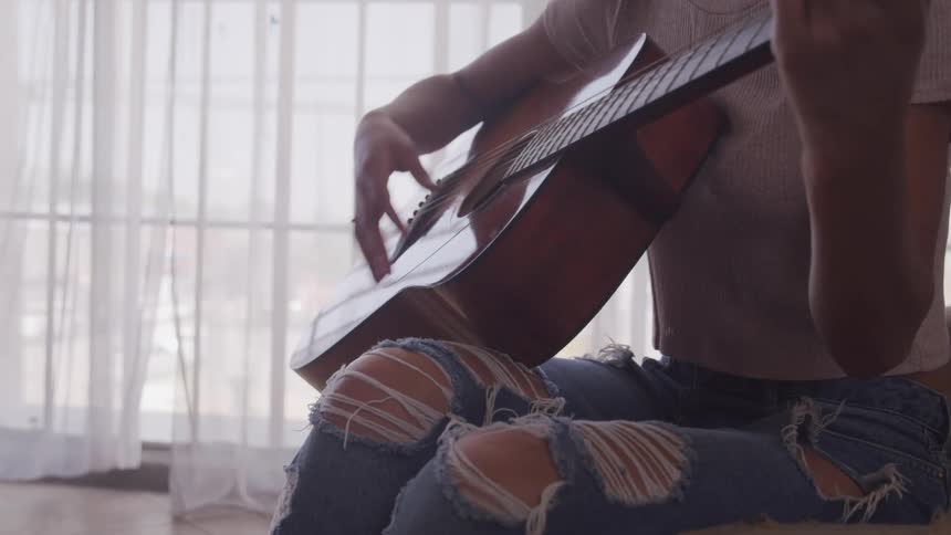 A woman sitting in front of a window at home plays the guitar