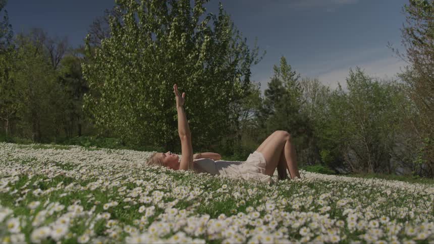 A teenage girl lying in a field of flower raises her arm towards the sky