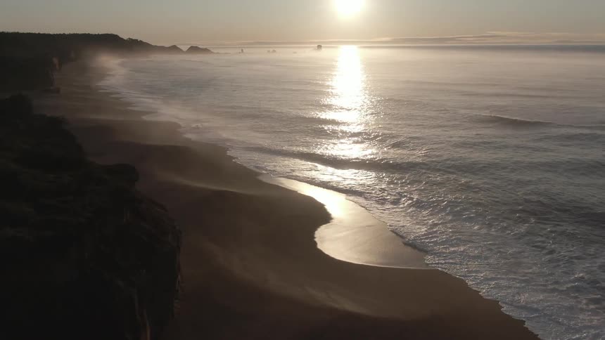 Ocean waves wash into a beach below a stone cliff during the day