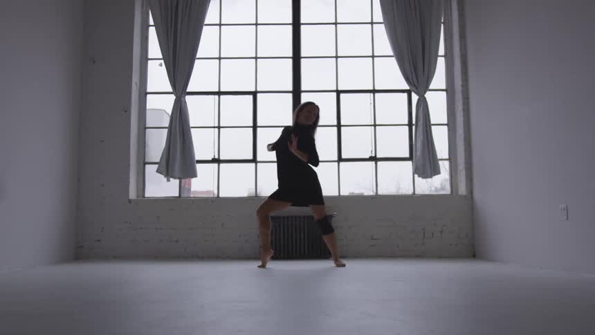 A woman in a black dress dancing in a room with a window in the background