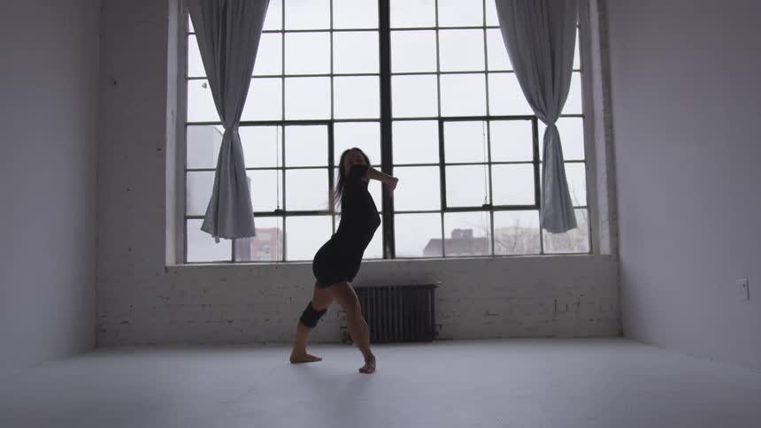 A woman dances in front of a large window in a white room