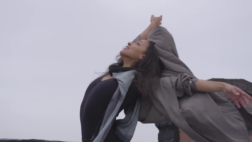 A woman stands outside as snow lightly falls and leans back with her arms raised in a dance pose