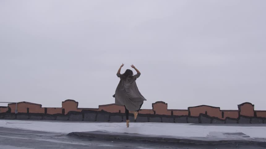 A young ballerina dances on a rooftop as snow falls around her from the cloudy sky above