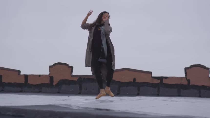 A young woman performs ballet moves on a rooftop as snow falls around her