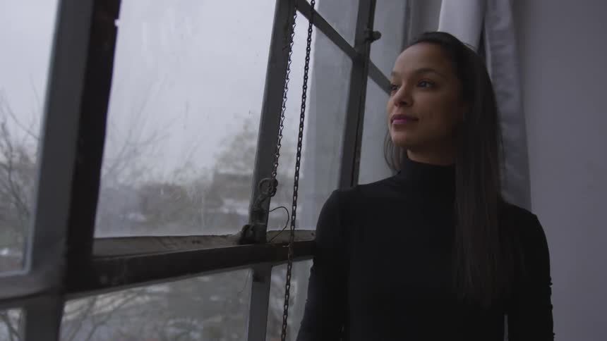 A young black woman looks out of a large window contemplatively on an autumn day