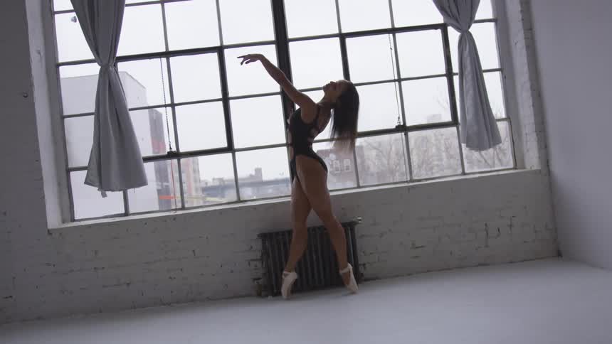 A female ballet dancer stands on the tips of her toes in front of a large window