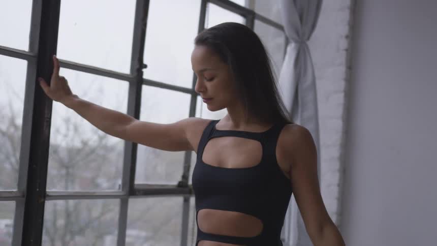 Young fit female dancer stands near a window in a studio on the city