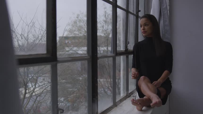 A woman in a black turtleneck dress sits in a large window with her legs up and looks outside at barren trees