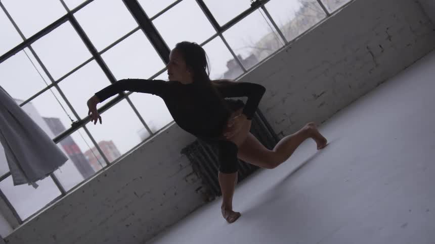 A woman performs a dance maneuver in front of laugh window in an old building