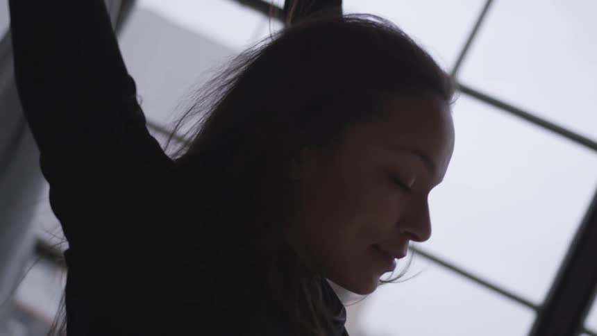 A young woman performs dance maneuvers in from of a set of large windows