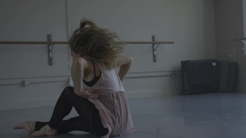 A dancer in a studio sits up from a laying position and moves her arms up and out