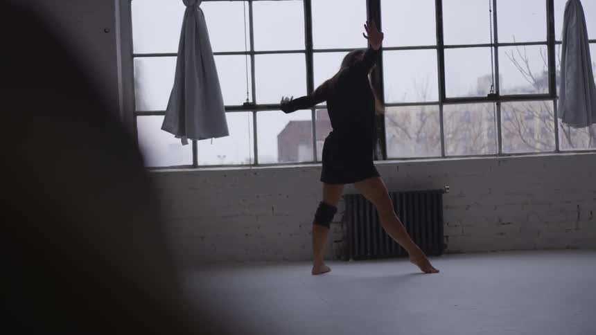 Woman dances in wide open room near window looking out onto buildings