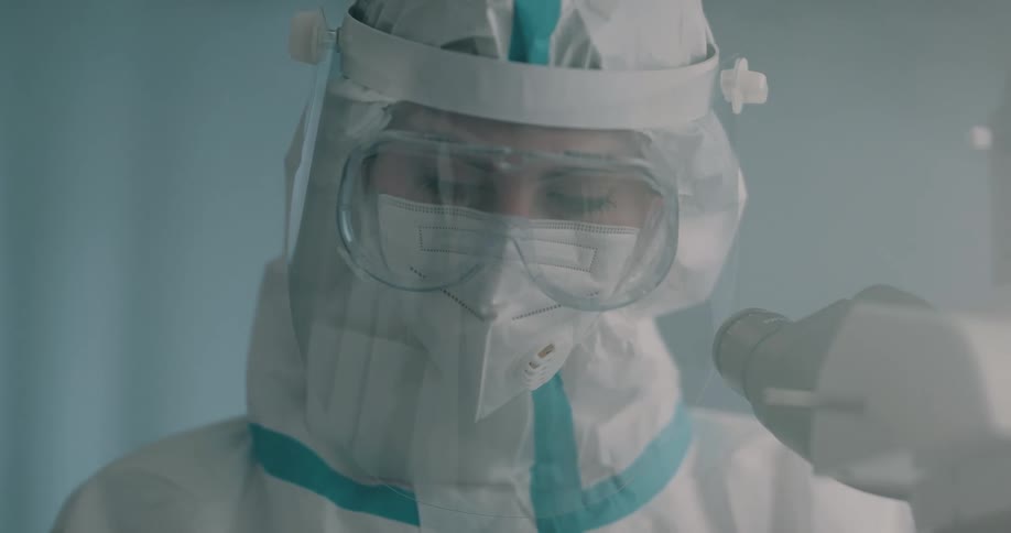 Woman dressed in full protective gear including face shield and face mask works with slide near microscope