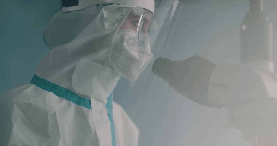 A woman in a lab wearing protective clothing and a face shield and googles looks through a microscope