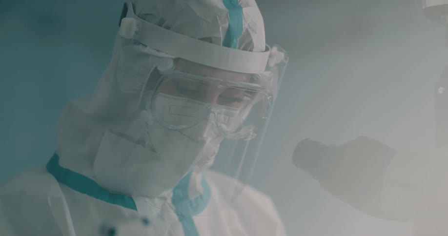 A person with a face shield and goggles in a lab next to a microscope