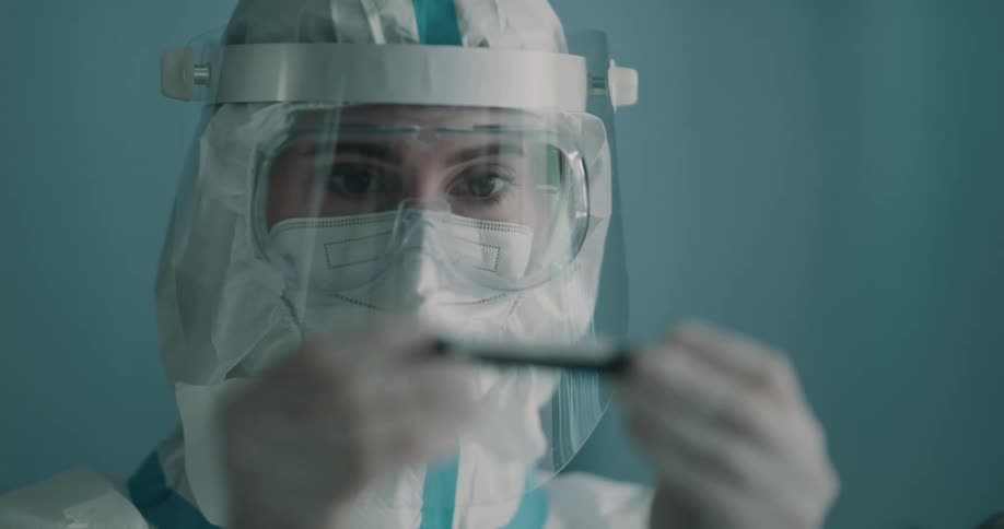 A woman in a hazmat suit looks at a tube of blood in a test tube