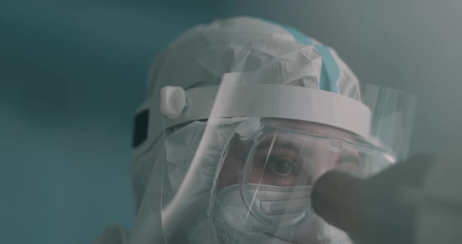 A female wearing a large amount of personal protective equipment looks into the lenses of a microscope