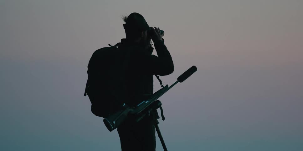 Hunter carrying a rifle looks through binoculars at the horizon